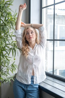 Pencerenin yanında beyaz gömlek ve kot pantolon giyen güzel sarışın genç bir kadın var. Arka planda yeşil bitkiler ve penceresi olan bir kızın sabah portresi.