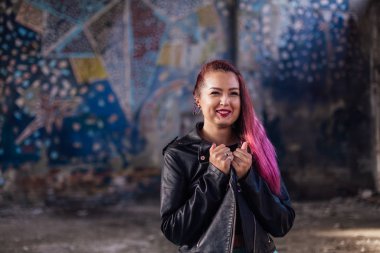 Deri ceketli ve mavi etekli pembe saçlı genç kız yıkılmış binanın içinde yıkıntılarla çevrili duruyor. Arka planda eski mozaik duvarın yanında duran kadın..