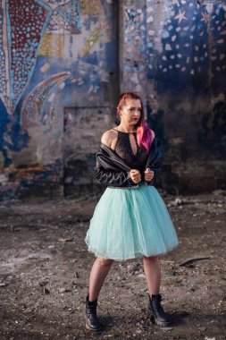 Deri ceketli ve mavi etekli pembe saçlı genç kız yıkılmış binanın içinde yıkıntılarla çevrili duruyor. Arka planda eski mozaik duvarın yanında duran kadın..