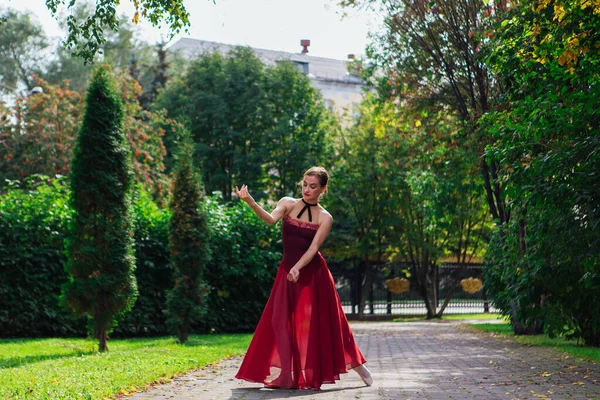 Woman ballerina in red ballet dress dancing in pointe shoes in autumn park. Ballerina standing in beautiful ballet pose