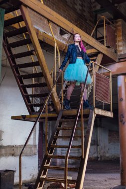 Pembe saçlı, deri ceketli ve mavi etekli bir kız çökmüş binanın içindeki paslı merdivenlerde dikiliyor. Etrafı harabelerle çevrili.