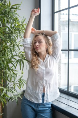 Pencerenin yanında beyaz gömlek ve kot pantolon giyen güzel sarışın genç bir kadın var. Arka planda yeşil bitkiler ve penceresi olan bir kızın sabah portresi.