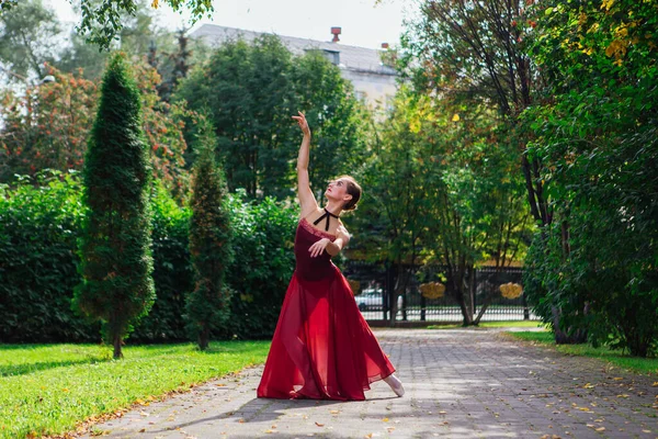 Woman ballerina in red ballet dress dancing in pointe shoes in autumn park. Ballerina standing in beautiful ballet pose