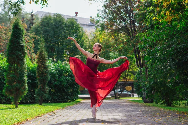 Woman ballerina in red ballet dress dancing in pointe shoes in autumn park. Ballerina standing in beautiful ballet pose