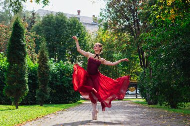 Sonbahar parkında kırmızı bale elbiseli kadın balerin, sivri uçlu ayakkabılarla dans ediyor. Balerin güzel bir bale pozisyonunda duruyor.