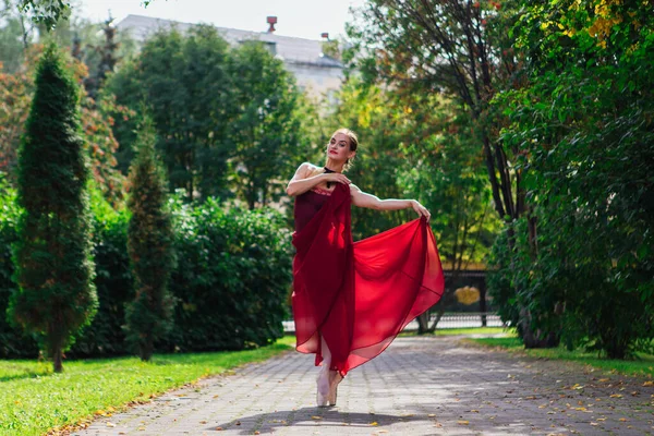 stock image Woman ballerina in red ballet dress dancing in pointe shoes in autumn park. Ballerina standing in beautiful ballet pose