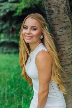 Güzel beyaz elbiseler giyen çekici bir sarışının bahar portresi. Parktaki Rowan ağacının yanında çimlerin üzerinde duruyor..