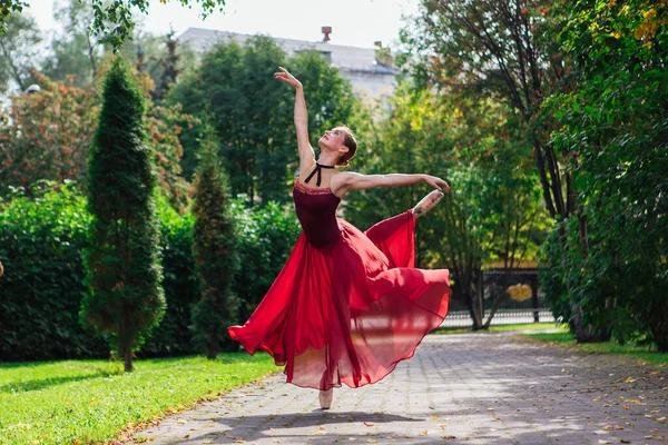 Woman ballerina in red ballet dress dancing in pointe shoes in autumn park.