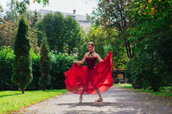 Woman ballerina in red ballet dress dancing in pointe shoes in autumn park. Ballerina standing in beautiful ballet pose