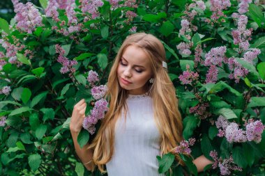 Güzel beyaz elbise giyen büyüleyici bir sarışın kadının bahar portresi çiçek açan mor leylak çalı yanında duran.