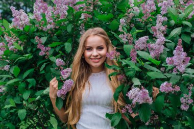 Güzel beyaz elbise giyen büyüleyici bir sarışın kadının bahar portresi çiçek açan mor leylak çalı yanında duran.