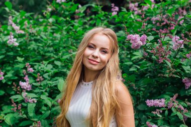 Güzel beyaz elbise giyen büyüleyici bir sarışın kadının bahar portresi çiçek açan mor leylak çalı yanında duran.