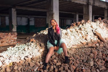 Pembe saçlı genç bir kız çöken bir binada, tuğlaların üzerinde oturuyor.