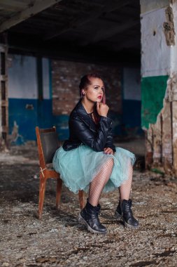 Pembe saçlı, deri ceketli ve mavi etekli genç bir kız yıkılmış binanın içindeki eski sandalyede oturuyor..