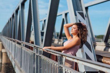 Gün batımında nehrin üzerindeki eski paslı nakliye köprüsünde poz veren genç ve güzel bir kadının ılık yaz portresi..