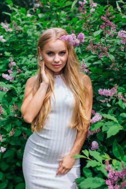 Güzel beyaz elbise giyen büyüleyici bir sarışın kadının bahar portresi çiçek açan mor leylak çalı yanında duran.