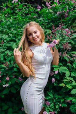 Güzel beyaz elbise giyen büyüleyici bir sarışın kadının bahar portresi çiçek açan mor leylak çalı yanında duran.