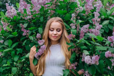 Güzel beyaz elbise giyen büyüleyici bir sarışın kadının bahar portresi çiçek açan mor leylak çalı yanında duran.