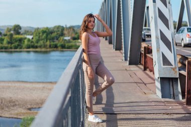 Gün batımında nehrin üzerindeki eski paslı nakliye köprüsünde poz veren genç ve güzel bir kadının ılık yaz portresi..