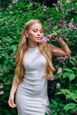 Güzel beyaz elbise giyen büyüleyici bir sarışın kadının bahar portresi çiçek açan mor leylak çalı yanında duran.