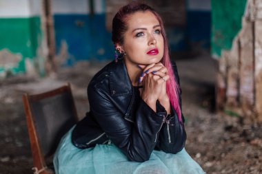 Pembe saçlı, deri ceketli ve mavi etekli genç bir kız yıkılmış binanın içindeki eski sandalyede oturuyor..