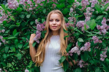 Güzel beyaz elbise giyen büyüleyici bir sarışın kadının bahar portresi çiçek açan mor leylak çalı yanında duran.