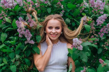 Güzel beyaz elbiseler giyen çekici bir sarışının bahar portresi yapraklarında saçı olan mor leylak çalılarının yanında duruyor.