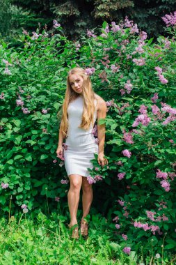 Güzel beyaz elbise giyen büyüleyici bir sarışın kadının bahar portresi çiçek açan mor leylak çalı yanında duran.