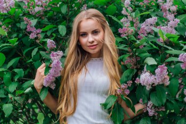 Güzel beyaz elbise giyen büyüleyici bir sarışın kadının bahar portresi çiçek açan mor leylak çalı yanında duran.
