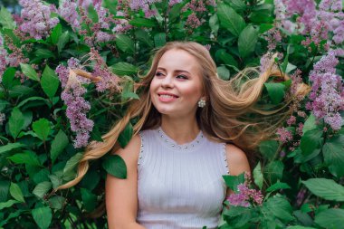 Güzel beyaz elbiseler giyen çekici bir sarışının bahar portresi yapraklarında saçı olan mor leylak çalılarının yanında duruyor.