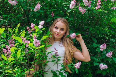 Güzel beyaz elbise giyen büyüleyici bir sarışın kadının bahar portresi çiçek açan mor leylak çalı yanında duran.