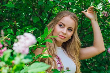 Güzel beyaz elbise giyen büyüleyici bir sarışın kadının bahar portresi çiçek açan mor leylak çalı yanında duran.