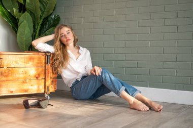 Genç, sarışın, beyaz gömlek ve kot pantolon giyen, yerde, tahta kutu ve yeşil bitkinin yanında çıplak ayakla oturan güzel bir kadın. Pencereden gün ışığında oturan bir kız. Boşluğu kopyala