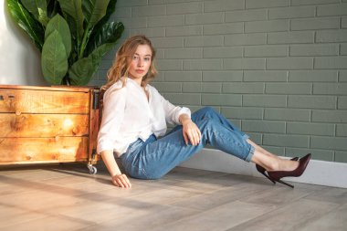 Beyaz gömlek ve kot pantolon giyen güzel sarışın kadın yerde, tahta kutu ve yeşil bitkinin yanında oturuyor. Pencereden gün ışığında oturan bir kız. Boşluğu kopyala