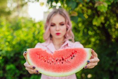 Güzel, gülümseyen, pembe saçlı genç bir kadın ellerinde tatlı bir karpuz tutuyor.