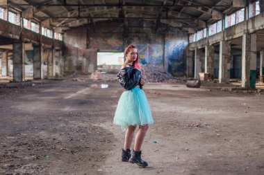 Deri ceketli ve mavi etekli pembe saçlı bir kızın portresi çökmüş binanın içinde yıkıntılarla çevrili duruyor.