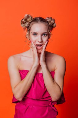 Stüdyoda turuncu arka planda kırmızı elbiseli kız. Sarışın kız, iki saç düğümlü. Elleriyle yavrularını tutuyor. Şaşkınlık gösteriyor.