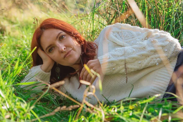 Morning portrait of a beautiful happy red woman with red hair laying in grass in white sweater