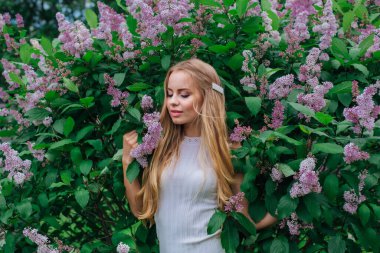 Güzel beyaz elbise giyen büyüleyici bir sarışın kadının bahar portresi çiçek açan mor leylak çalı yanında duran.