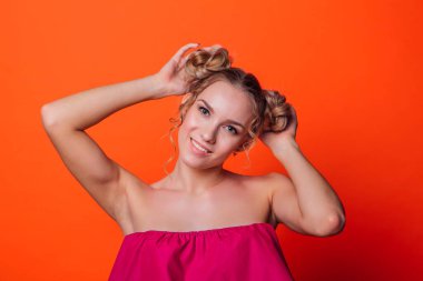 Stüdyoda turuncu arka planda kırmızı elbiseli kız. Sarışın, iki saç düğümlü, el ele tutuşmuş, kameraya bakıyor ve gülümsüyor..