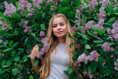 Güzel beyaz elbise giyen büyüleyici bir sarışın kadının bahar portresi çiçek açan mor leylak çalı yanında duran.