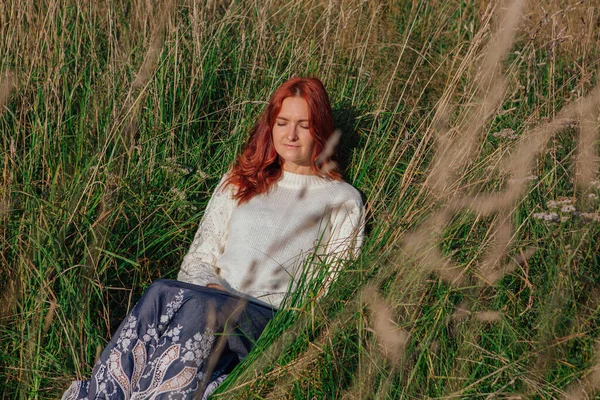Morning portrait of a beautiful happy red woman with red hair sitting in grass in white sweater