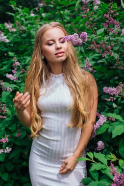 Güzel beyaz elbise giyen büyüleyici bir sarışın kadının bahar portresi çiçek açan mor leylak çalı yanında duran.