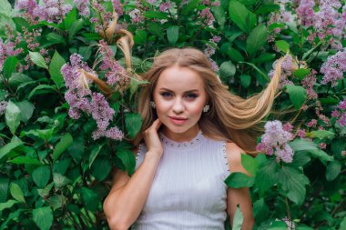 Güzel beyaz elbiseler giyen çekici bir sarışının bahar portresi yapraklarında saçı olan mor leylak çalılarının yanında duruyor.
