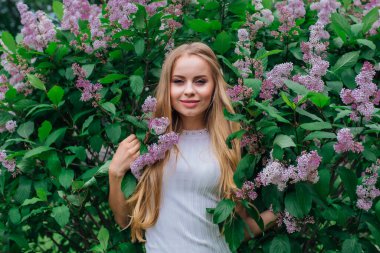 Güzel beyaz elbise giyen büyüleyici bir sarışın kadının bahar portresi çiçek açan mor leylak çalı yanında duran.
