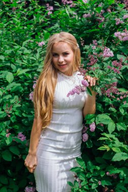 Güzel beyaz elbise giyen büyüleyici bir sarışın kadının bahar portresi çiçek açan mor leylak çalı yanında duran.
