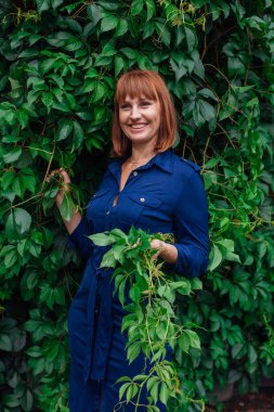 Güzel orta yaşlı bir kadının portresi. Makyajlı ve kızıl saçlı. Yaban üzüm yapraklarının yanında duruyor..