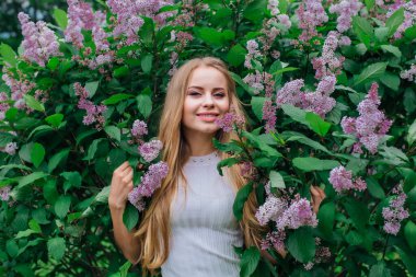 Güzel beyaz elbise giyen büyüleyici bir sarışın kadının bahar portresi çiçek açan mor leylak çalı yanında duran.