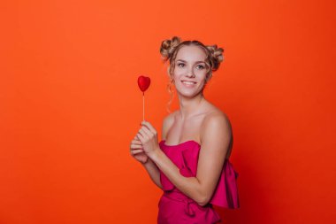 Stüdyoda turuncu arka planda kırmızı elbiseli kız. Sarışın, iki saç düğümlü, kırmızı kalbi çubukta tutan ve gülümseyen kız. Boşluğu kopyala.