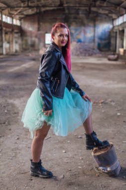 Deri ceketli ve mavi etekli pembe saçlı bir kızın portresi çökmüş binanın içinde yıkıntılarla çevrili duruyor.